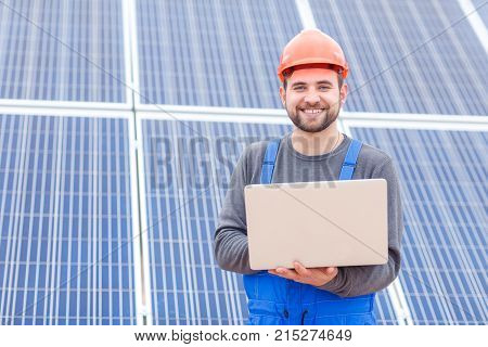 A happy worker at the solar battery station is dressed in a uniform and helmet and is holding a gray modern laptop. Outdoors.