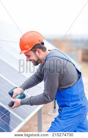 A happy worker at the solar battery station with a beard, dressed in a blue uniform and an orange helmet drills a solar battery panel. Outdoors.