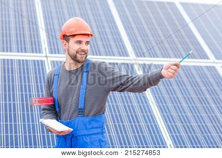 A young bearded worker at the solar battery station in blue uniform shows his hand somewhere and holds a notebook, a pencil and waterpas in his hands. Outdoors.
