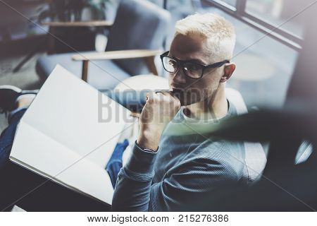 Portrait handsome young man wearing glasses casual clothes holding book hands.Man sitting in vintage armchair modern loft studio reading book. Blurred background.Horizontal