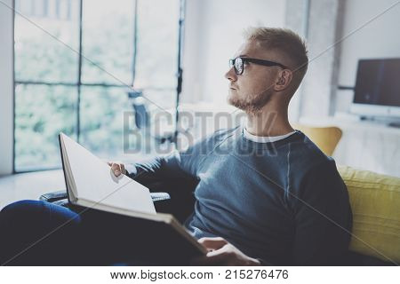 Portrait handsome young man wearing glasses casual clothes holding book hands.Man sitting in vintage armchair modern loft studio reading book. Blurred background.Horizontal