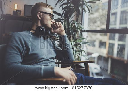 Pensive young man wearing glasses casual clothes.Man sitting in vintage armchair modern loft studio and relaxing whith headphone music. Blurred background.Horizontal