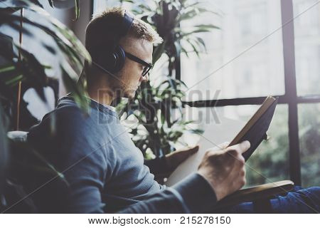 Attractive young man wearing glasses casual clothes.Man sitting in vintage armchair modern loft studio, reading book and relaxing whith headphone music. Blurred background.Horizontal