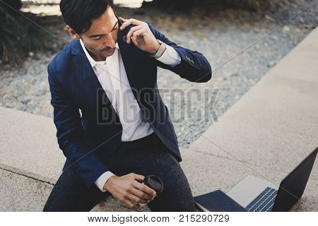 Businessman using contemporary smartphone taking rest outside the office during work day.Holding hands take away coffee cup.Blurred background. Horizontal