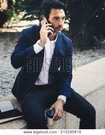 Pensive elegant businessman using contemporary smartphone while taking rest during work day.Holding hands take away coffee cup.Blurred background. Vertical