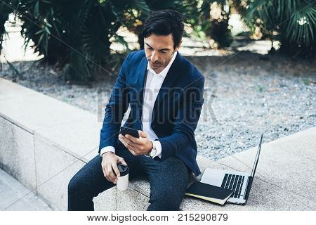 Attractive smiling businessman using contemporary smartphone taking rest during work day.Holding hands take away coffee cup.Blurred background. Horizontal.Cropped