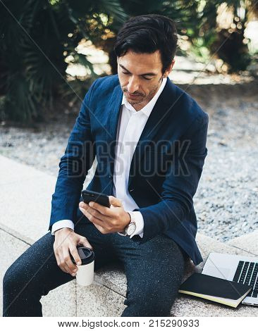 Attractive smiling businessman using contemporary smartphone taking rest during work day.Holding hands take away coffee cup.Blurred background. Horizontal.Cropped