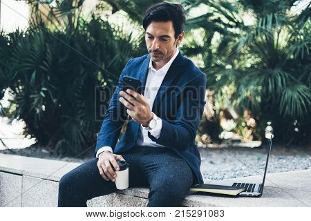 Attractive smiling businessman using contemporary smartphone taking rest during work day.Holding hands take away coffee cup.Blurred background. Horizontal
