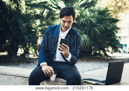 Attractive smiling businessman making video conversation with partners by mobile phone.Man using contemporary smartphone sitting outside.Blurred background. Horizontal