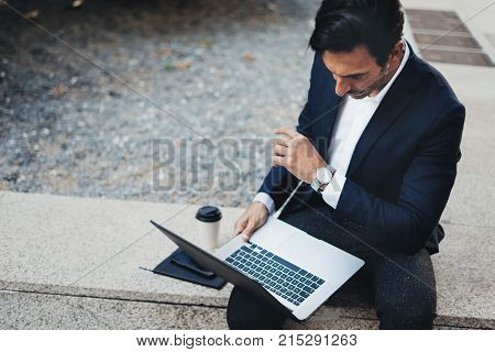Attractive elegant businessman using contemporary notebook and smartphone sitting outside.Blurred background. Horizontal