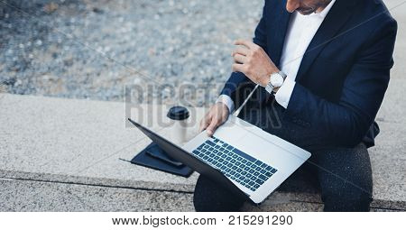 Attractive elegant businessman using contemporary notebook and smartphone sitting outside.Blurred background. Horizontal.Cropped