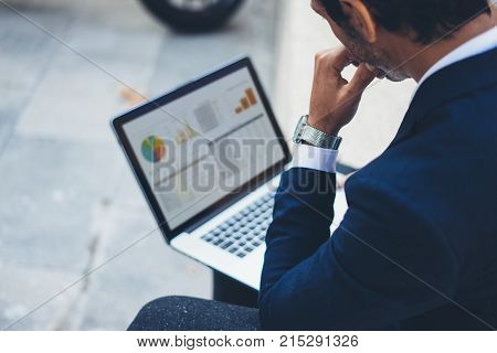 Attractive elegant businessman using contemporary notebook sitting outside the office.Graphs and diagramm on laptop screen.Blurred background. Horizontal