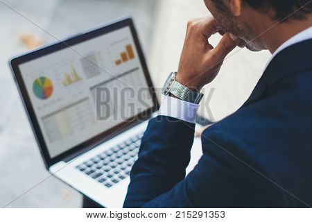 Attractive elegant businessman using contemporary notebook sitting outside the office.Graphs and diagramm on laptop screen.Blurred background. Horizontal.Cropped