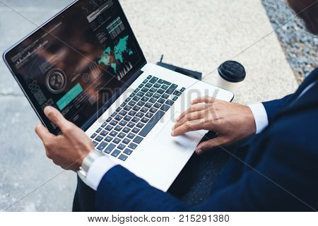 Attractive elegant businessman using contemporary notebook sitting outside the office.Graphs and diagramm on laptop display.Blurred background. Horizontal.Cropped