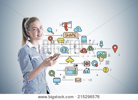 Portrait of a smiling young businesswoman wearing a blue shirt and holding a smartphone. A gray wall background with a social media sketch