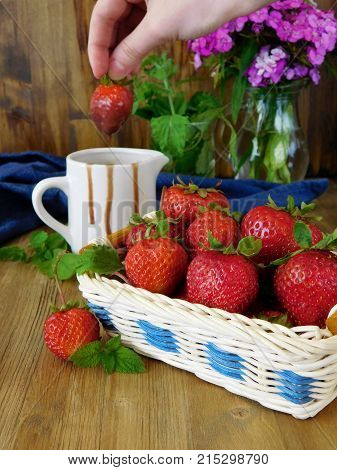 Strawberries in a wicker basket and sauce in a ceramic jug in the background. Female hand is dipping the berry into the chocolate