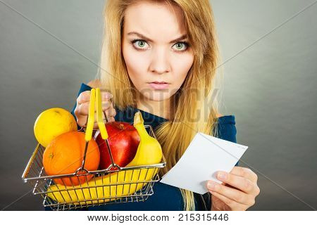 Worried woman holding shopping basket with fruits looking at bill receipt being scared of huge prices