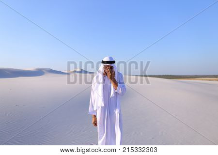 Cheerful Muslim guy walks along white sand of wide desert on warm summer evening. Arab young man in national clothes dances on spot and laughs giggly, adjusting hands with harness in wind. Swarthy, handsome Muslim with short dark hair dressed in kandura, 