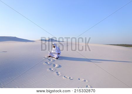 Beautiful Emirate Sheikh businessman studying project of future construction of complex, standing in middle of bottomless desert with white clean sand on clear warm day against blue sky. Swarthy Muslim with short dark hair dressed in kandura, long, spacio