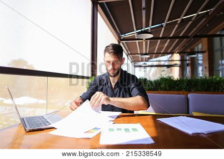Placeman sorting papers on desk near laptop. Strong male hands with watch near window. Concept of stopping document clutter on workplace.