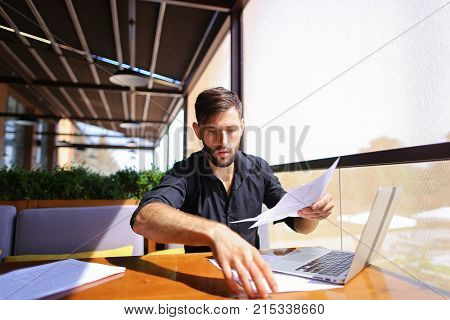 Placeman sorting papers on desk near laptop. Strong male hands with watch near window. Concept of stopping document clutter on workplace.