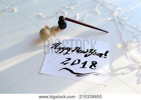 Christmas card with festive inscription Happy New Year, painted with black calligraphic font using carcass and fountain pen, which lies on table with Christmas garland in art workshop. Concept of beautiful writing, calligraphy and lettering, materials and