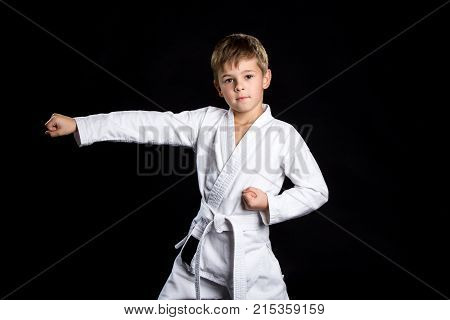 Straight karate arm hit. Kid in brand new kimono looking on the camera on isolated black background.