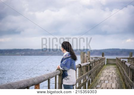 Woman on a bridge looking in the distance - Young woman standing alone on an old wooden bridge thinking and gazing the horizon in the afternoon over the Chiemsee lake in Bavaria Germany.
