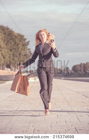 Vertical orange tonted shot of a happy beautiful blond woman running with paper bags and phone in hand along the promenade