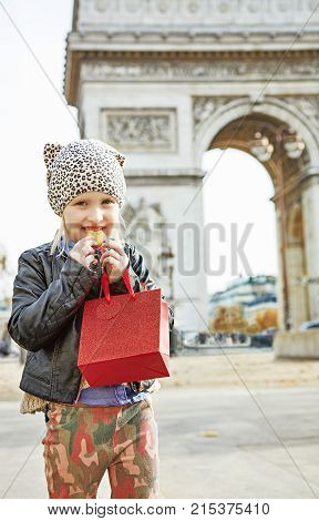 Stylish autumn in Paris. happy modern child with red present bag near Arc de Triomphe in Paris eating French macaroon