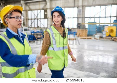Confident experienced forewoman walking with female colleague over factory workshop and explaining her work