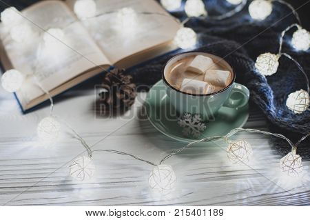 Cozy winter home. Cup of cocoa with marshmallows warm knitted sweater open book Christmas garland lump on a white wooden table. Atmosphere of a pleasant evening for reading.