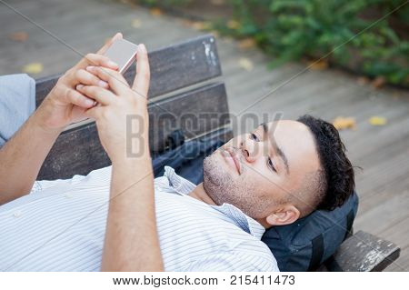 Closeup portrait of young relaxed student lying on bench in park with backpack under head and texting message to his girlfriend. Life balance concept