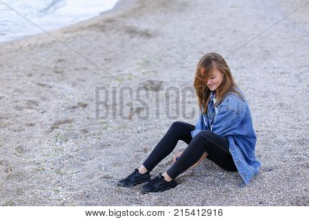 Dreamy girl relaxes and enjoys rest on seaside, throws head back and shakes hair, sitting on sandy beach on Black Sea shore on clear evening. Young European-looking woman with blond mid-length hair dressed in white jacket over which blue denim jacket, bla