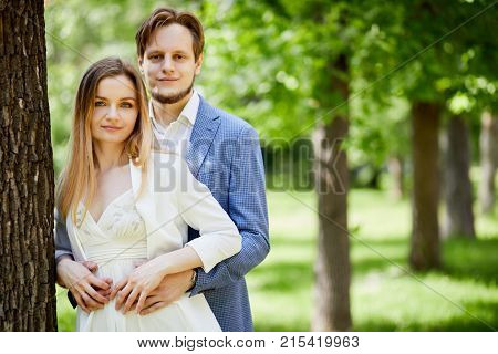 Young bearded man and woman stand ebracing at big tree trunk in park.