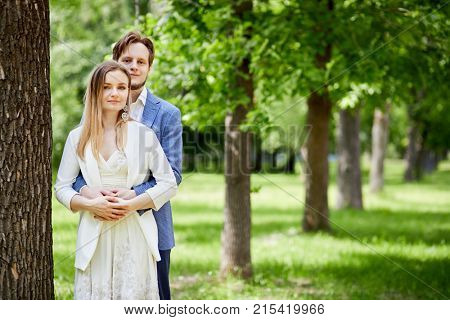 Young bearded man and woman stand ebracing at big tree trunk in summer park.