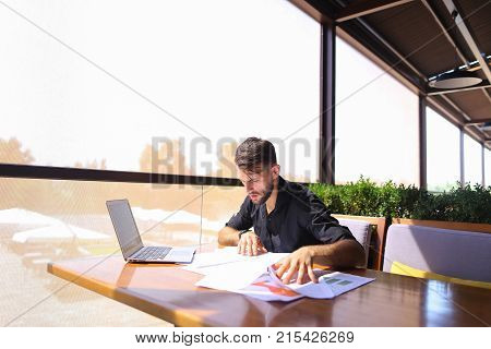 Placeman sorting papers on desk near laptop. Strong male hands with watch near window. Concept of stopping document clutter on workplace.