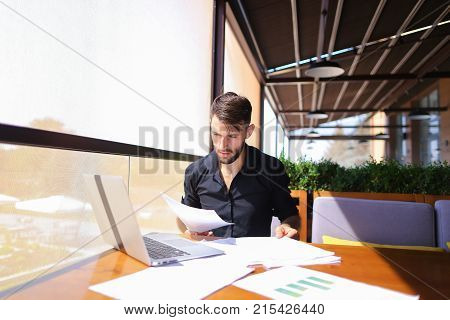 Placeman sorting papers on desk near laptop. Strong male hands with watch near window 