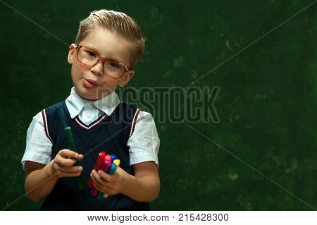 Cheerful little boy in glasses with fashionable styling in formal attire looks into the camera saying something and holding pens on dark green background
