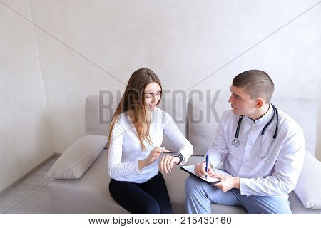 Experienced cardiologist doctor shows girl to client with iWatch on arm how to measure and monitor heart rate, gives recommendations and advice, woman listens attentively while sitting on couch on background of gray wall in hospital office during day. Eur