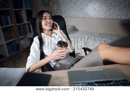 The girl is working late in the dark office with a laptop. Young beautiful businesswoman girl in office. A girl in a skirt has put her feet on the table and is talking on the phone in the office.