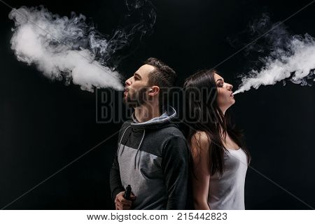 Couple vaping. Young man and woman blowing smoke at black studio background. Relationship and vape addiction concept with copy space