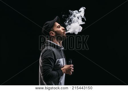 Young man vaping, studio shot. Bearded guy blowing a cloud of smoke on black background. Nicotine free smoking and vapor concept, copy space