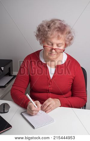 An elderly woman writes a letter to the white table.