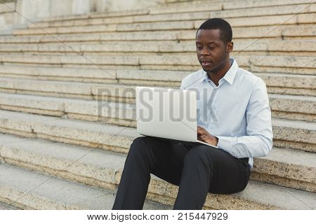 Serious black businessman working on laptop. African-american insuranse agent checking email on computer, sitting on the stairs outdoors in urban area