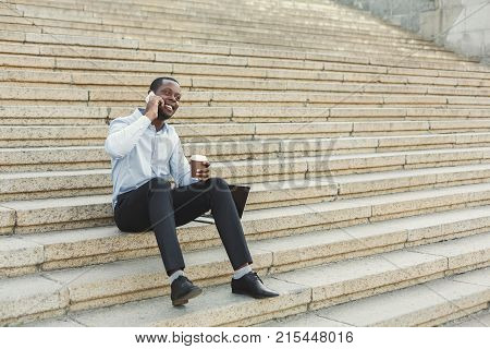 Happy black businessman working on laptop. African-american insuranse agent checking email on computer, talking on mobile, sitting on the stairs outdoors in urban area