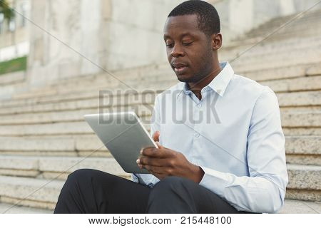 Serious black businessman working on tablet. African-american insuranse agent browsing news on digital device, sitting on the stairs outdoors in urban area