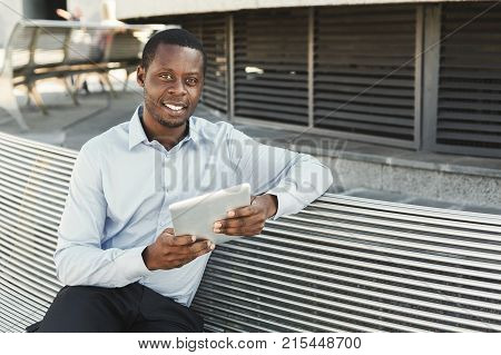 Happy black businessman working on tablet. African-american insuranse agent browsing news on digital device, sitting on the bench outdoors near modern office center in urban area cityscape