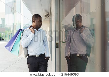 Happy black man with shopping bags walking along modern boutiques street. Successful business and shopping concept