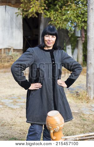 Smiling woman with black hair in ancient national clothes stands near the fence against the backdrop of rural farmstead. Ukrainian village. Ukraine